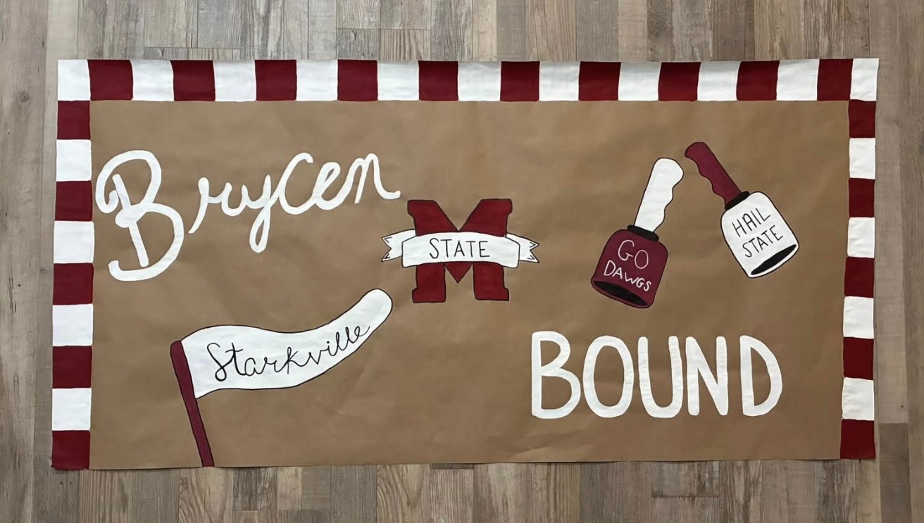Hand-painted kraft paper banner reading 'Bryçen — Starkville — Bound' with a maroon M State logo and checkered maroon and white border